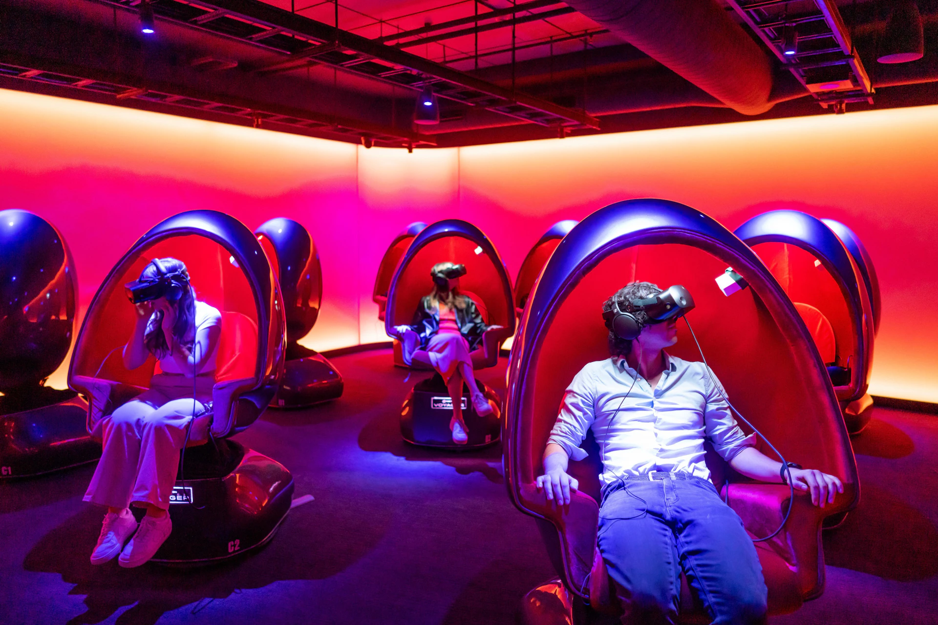 People seated in pod-like chairs, wearing virtual reality headsets, in a room with vibrant pink and purple lighting.