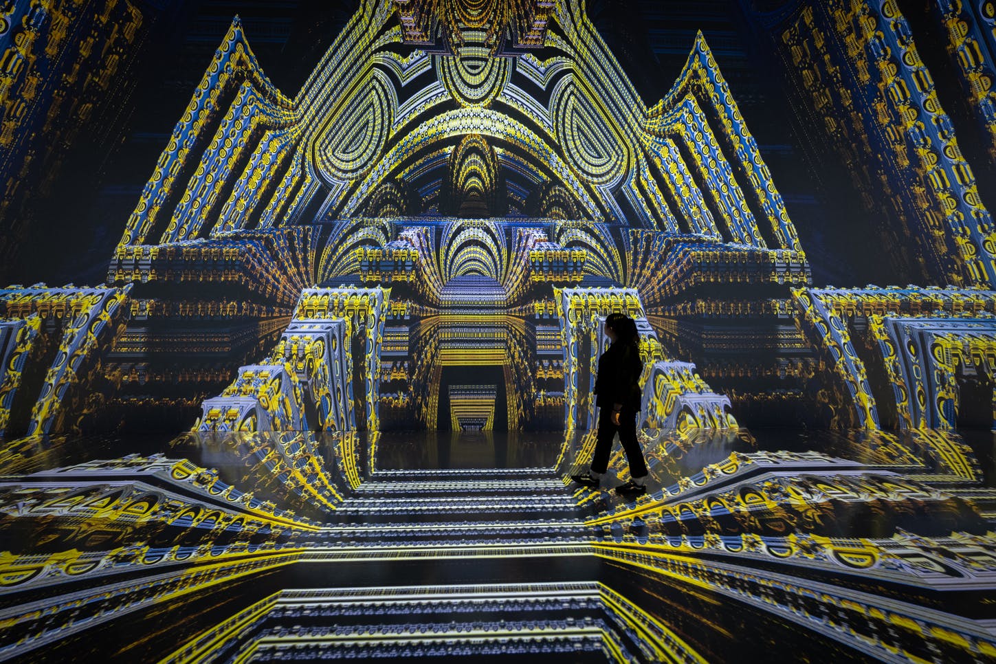 A person walking in a room with immersive, intricate yellow and blue digital projections resembling an ornate structure.