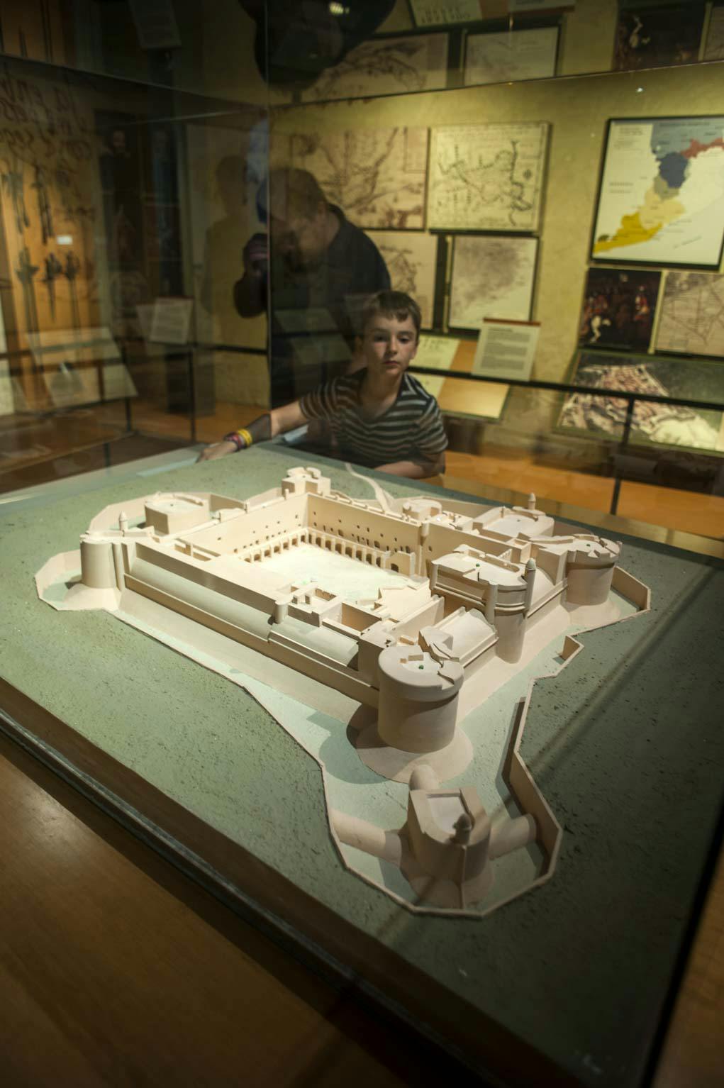 A child observes a scale model of a historical fort displayed in a glass case, with maps and diagrams on the wall behind.