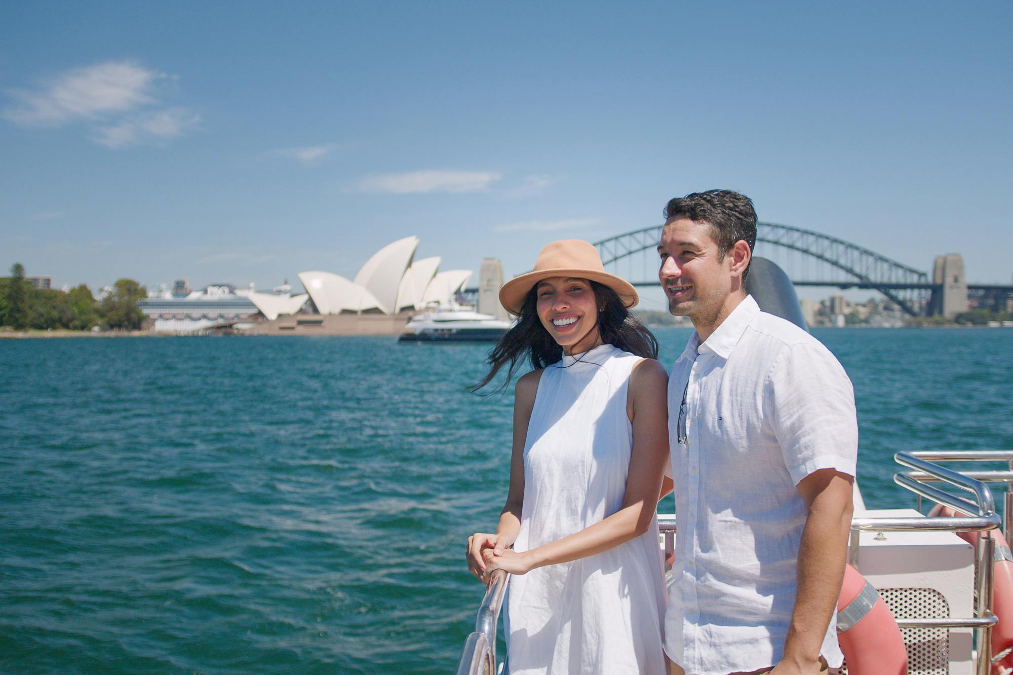 A couple on a boat with the Sydney Opera House and Harbour Bridge visible in the background on a sunny day.