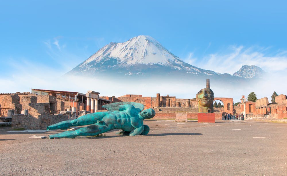A large blue statue lies on the ground in front of ancient ruins with a snow-capped mountain in the background.