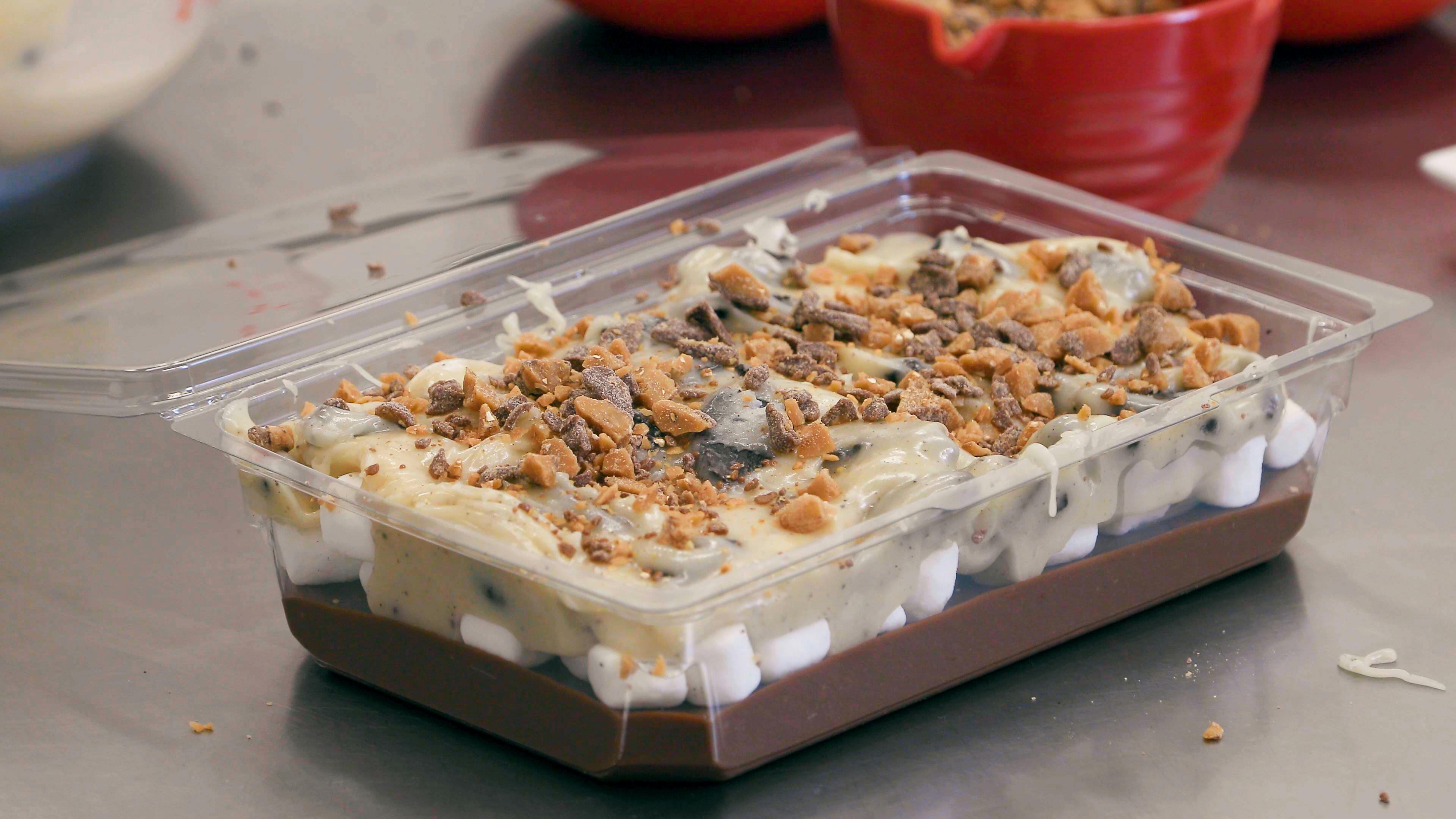 Een doorzichtige plastic schaal gevuld met een gelaagd dessert van chocolade, marshmallows, room en verkruimelde stukjes toffee bovenop.