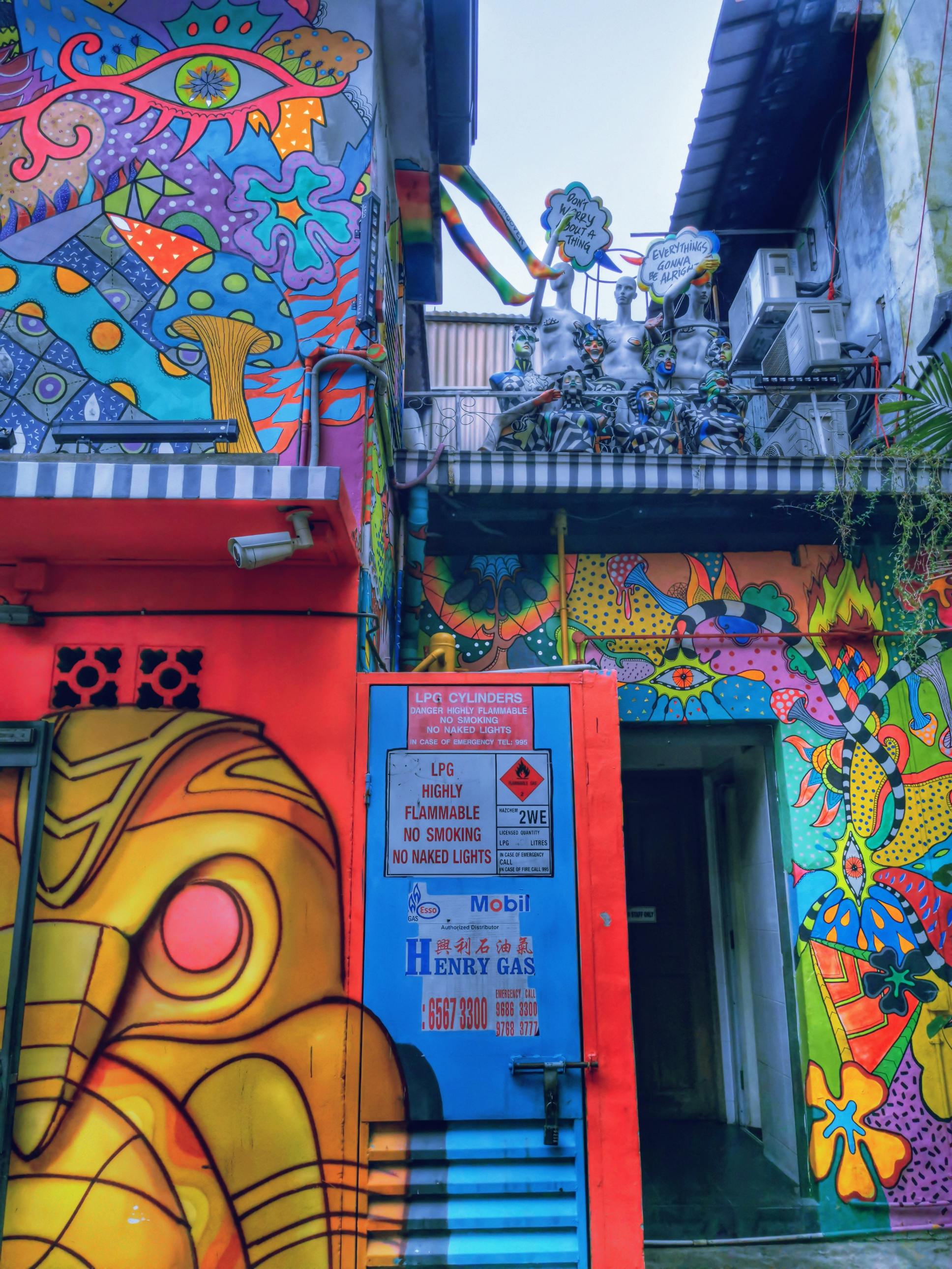 Colorful, mural-covered building exterior with psychedelic designs, a blue gas tank door, and various warning signs. Air conditioners and plants visible.