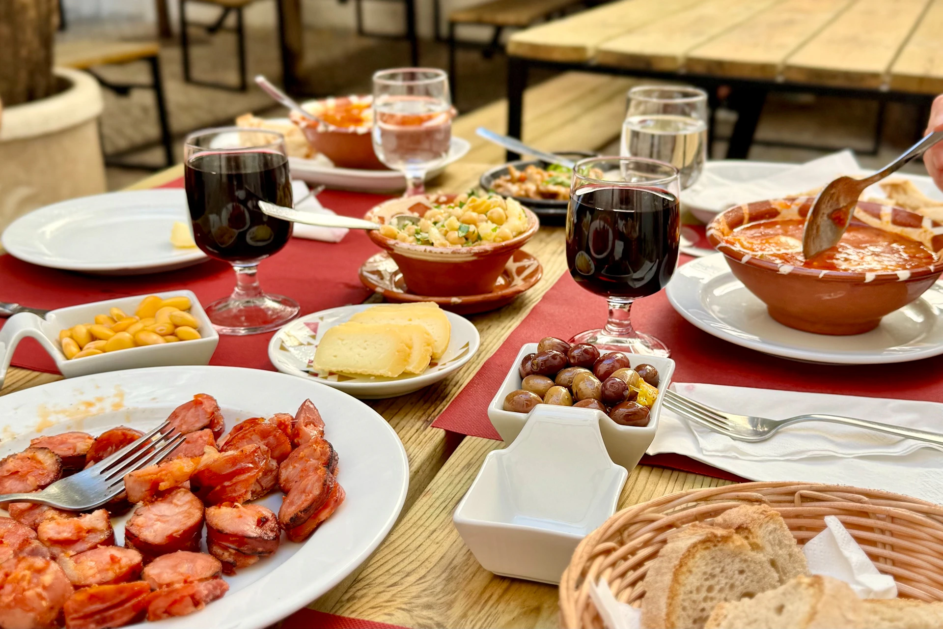 A table set with various dishes, including sausages, cheese, olives, salad, and glasses of red wine, in an outdoor dining area.
