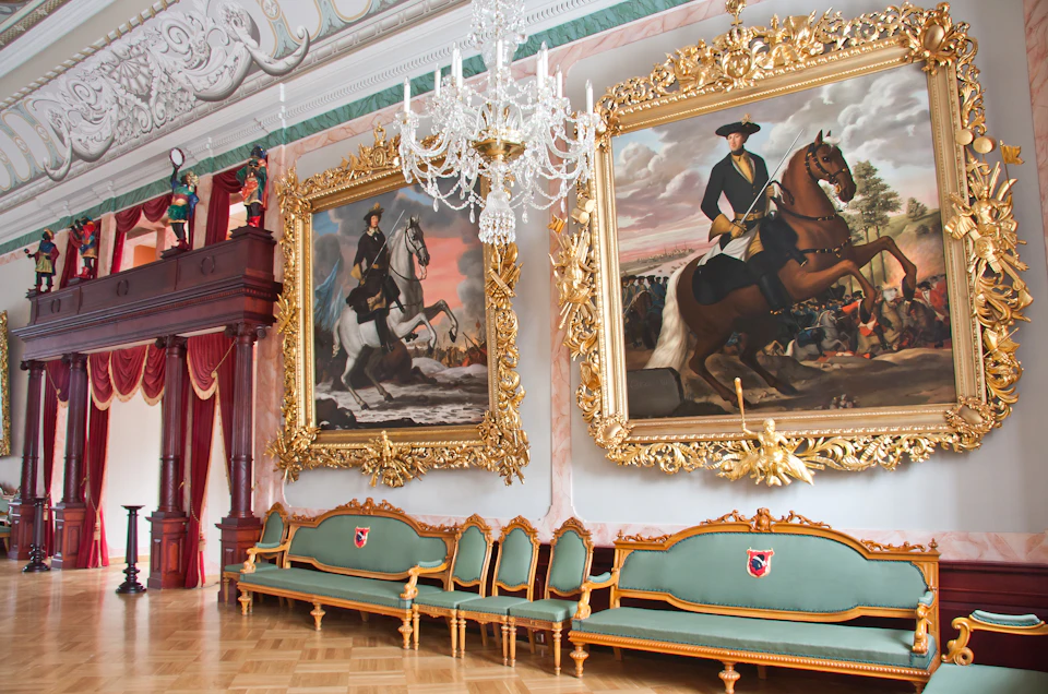 A grand room with two large, ornate-framed paintings of men on horses, green sofas, chairs with gold accents, and a chandelier.