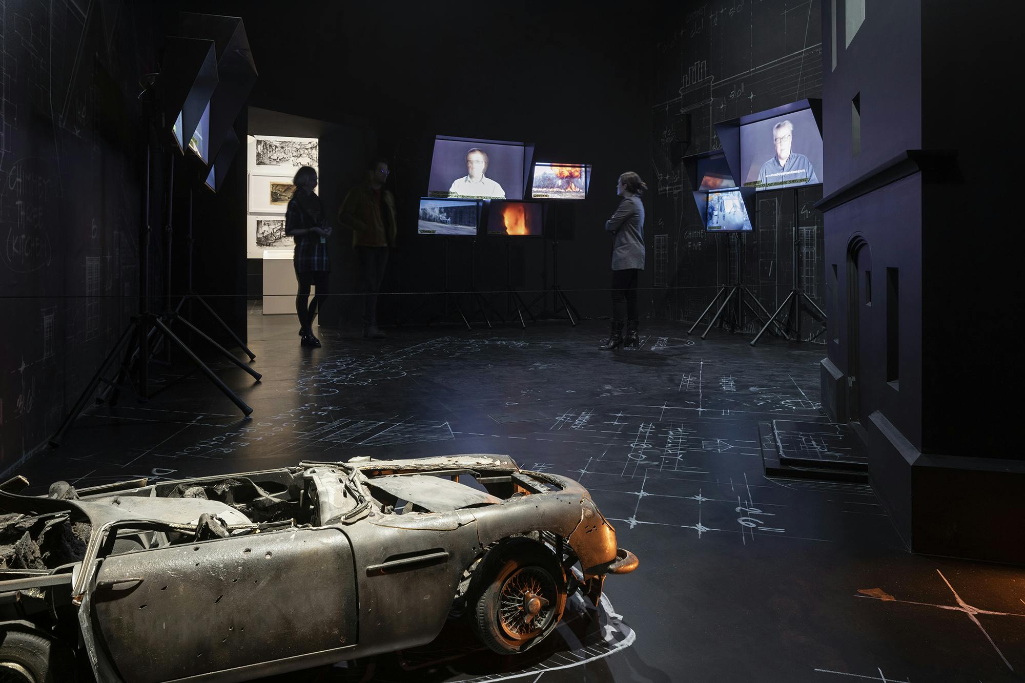 Σκοτεινό δωμάτιο που εμφανίζει πολλές οθόνες με ανθρώπους πάνω τους, πολλά άτομα παρατηρούν και ένα καμένο αυτοκίνητο σε πρώτο πλάνο.