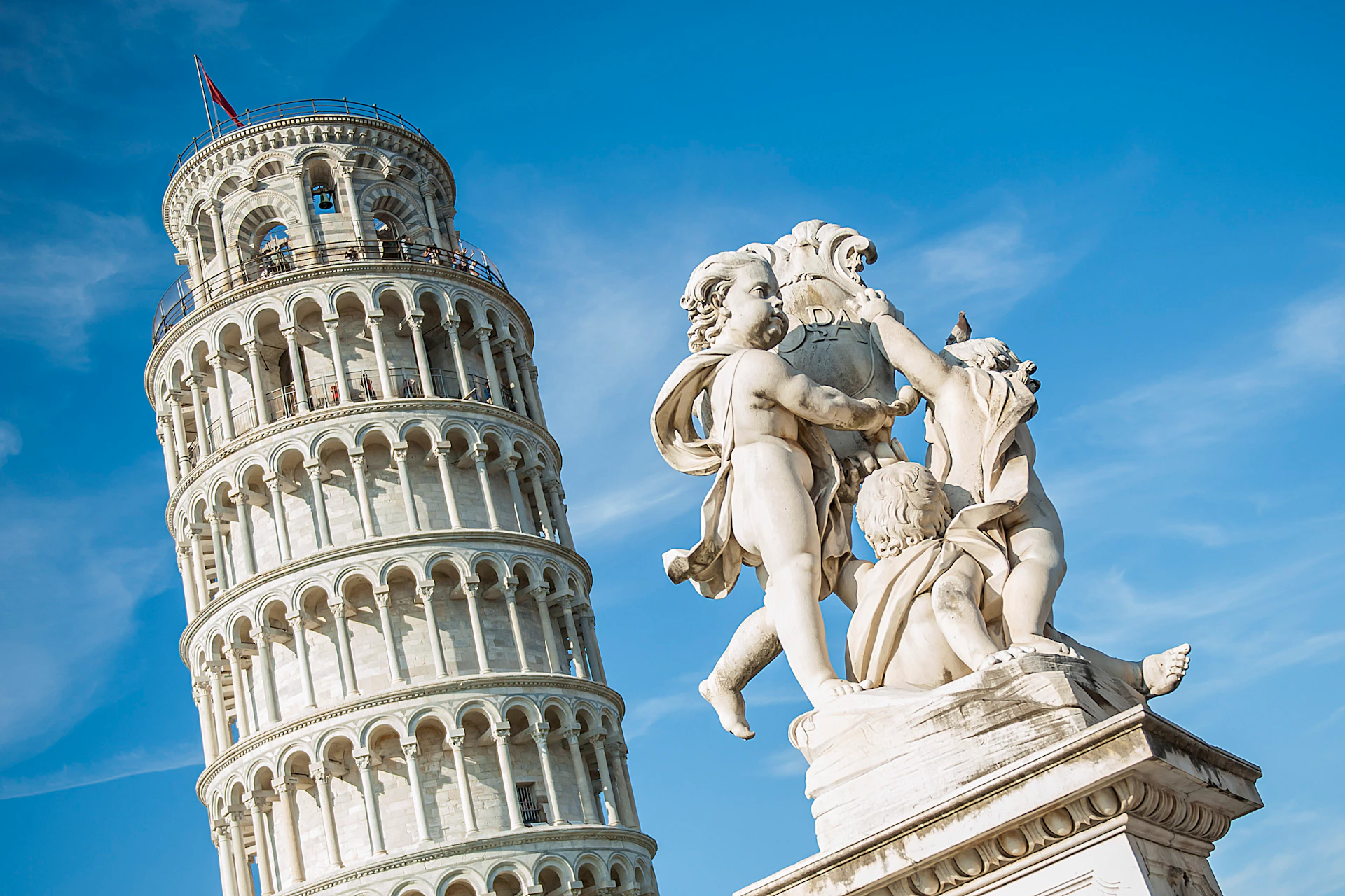 White marble statue of three cherubs in front of the Leaning Tower of Pisa under a clear blue sky.