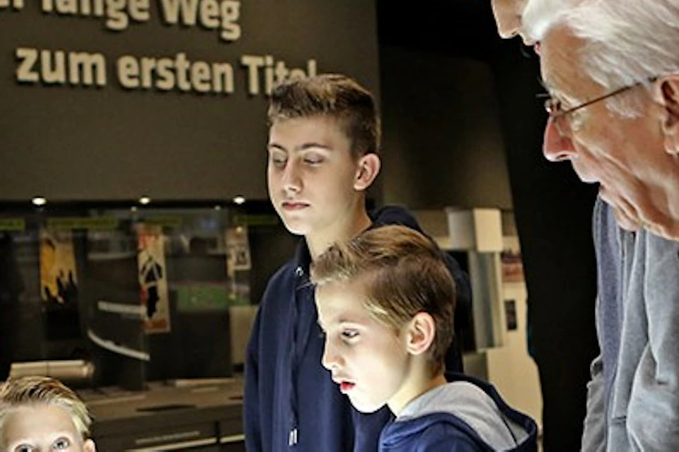 A group of boys and an elderly man examine an interactive display with buttons. German text on the wall reads "Der lange Weg zum ersten Titel."