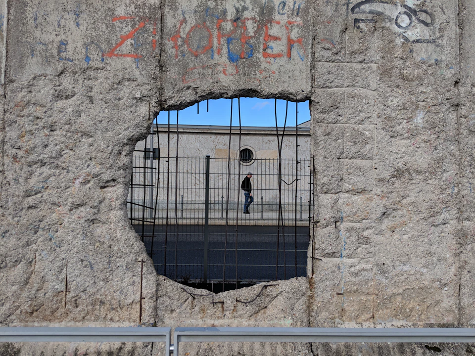 A man walks behind a concrete wall with a large hole, revealing wire and graffiti. A building is visible in the background.