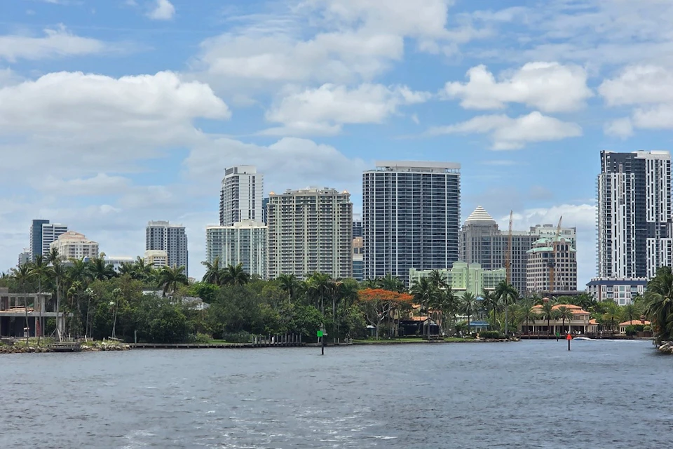 A city skyline with modern high-rise buildings, palm trees, and water in the foreground under a partly cloudy sky.