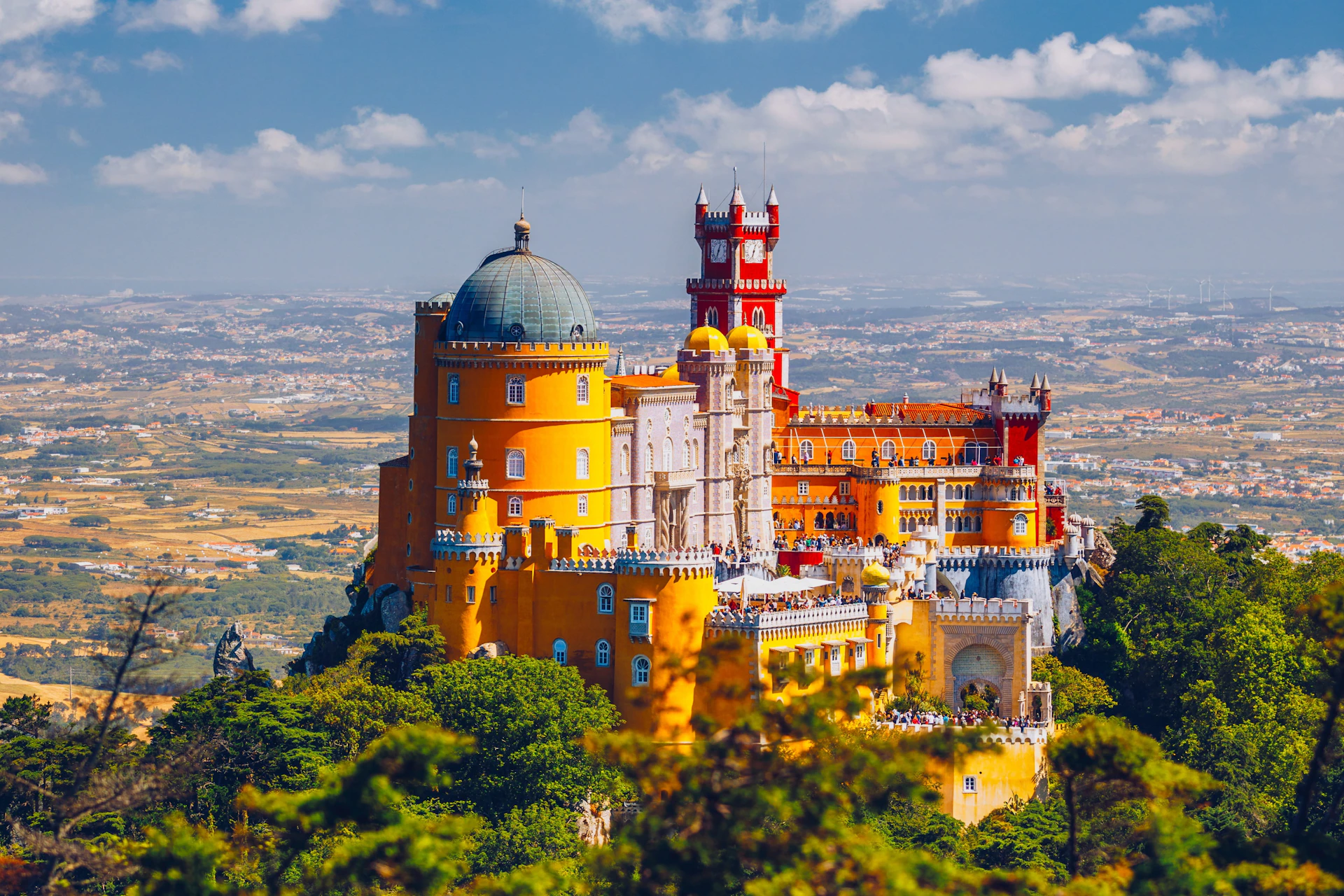 A colorful castle with yellow, red, and white sections on a hilltop surrounded by greenery under a blue sky.