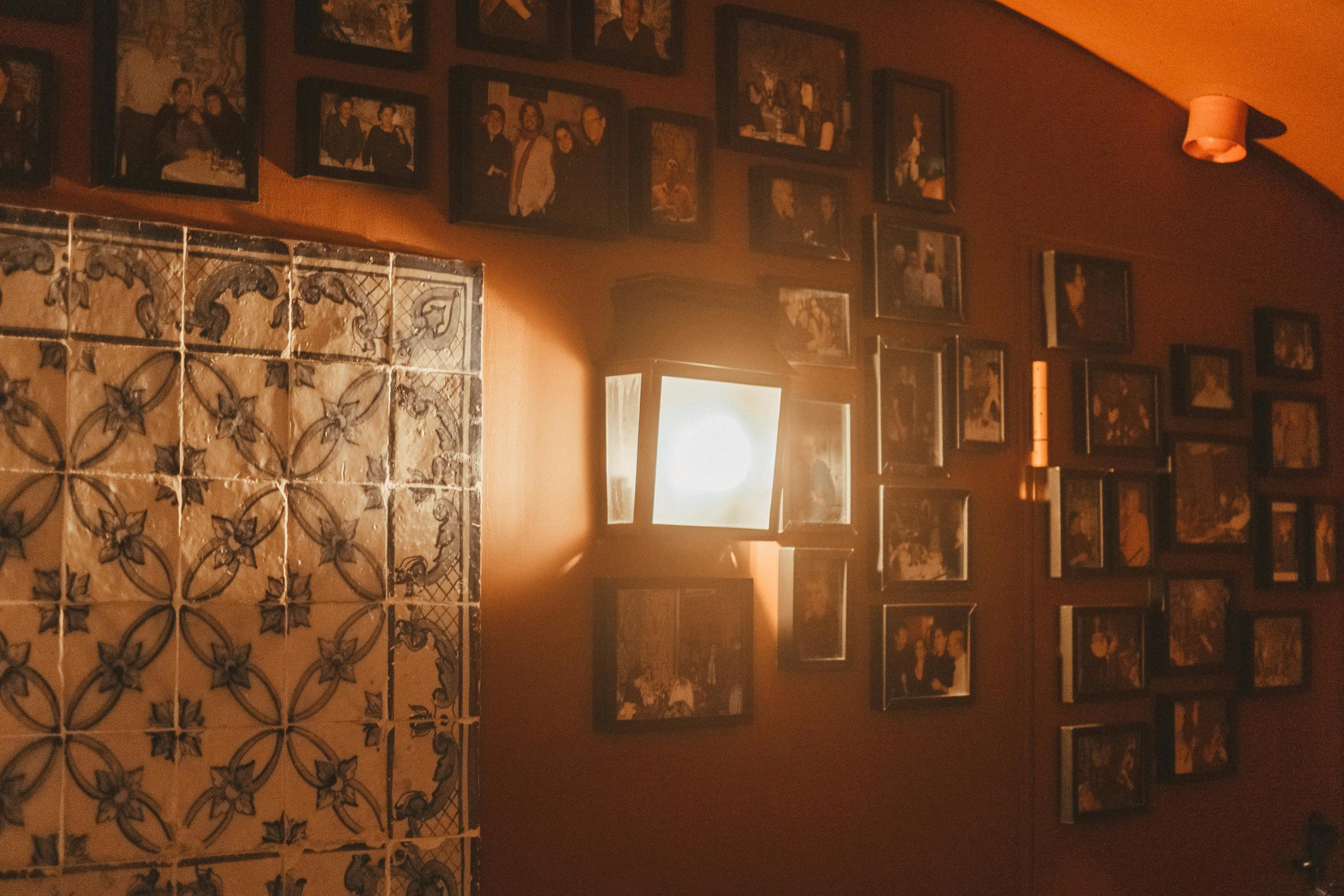 Ένα επιτοίχιο φωτιστικό φωτίζει κορνιζαρισμένες φωτογραφίες και διακοσμητικά πλακάκια σε έναν πορτοκαλί τοίχο.