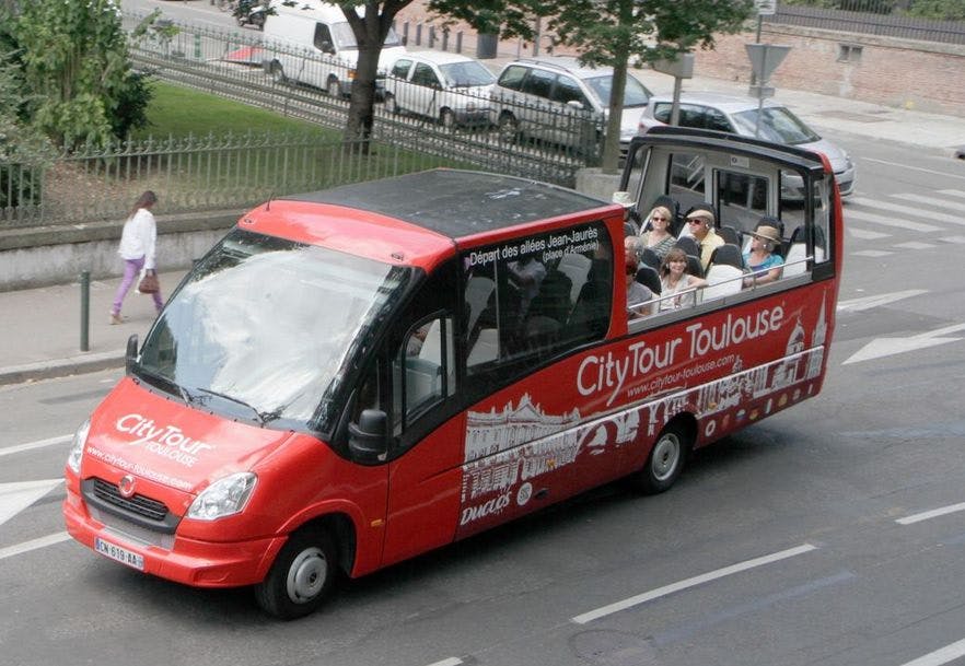 Ένα κόκκινο λεωφορείο City Tour Toulouse με επιβάτες που κάθονται στο επάνω ανοιχτό κατάστρωμα, οδηγώντας μέσα από έναν αστικό δρόμο.