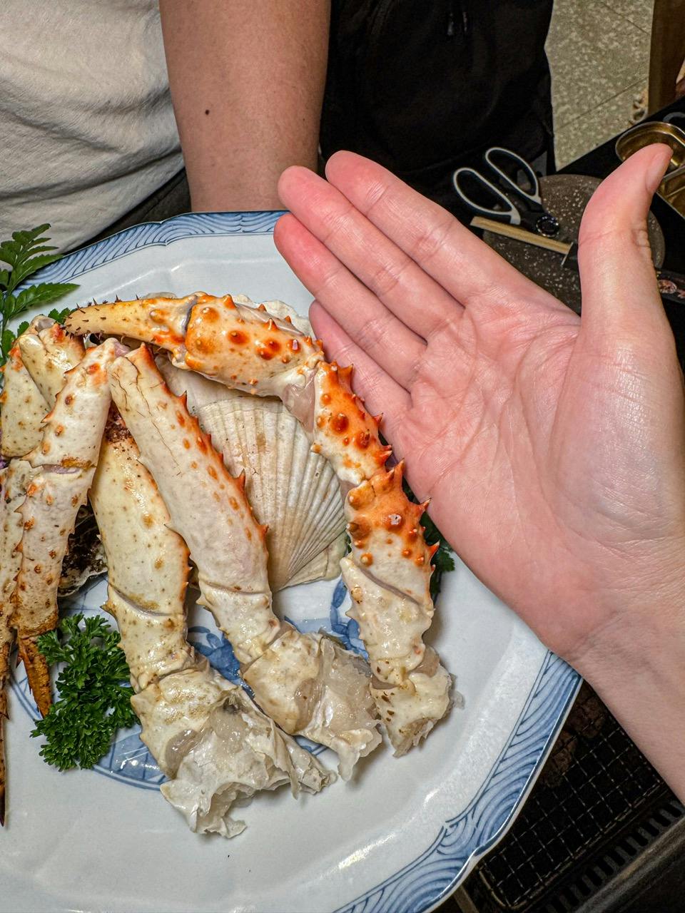 A hand held above a plate with large crab legs, a shell, and parsley garnish, showcasing the size of the crab legs.