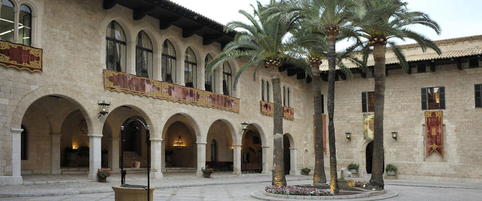 A stone courtyard with arched walkways, an upper balcony with decorations, palm trees, and potted plants.