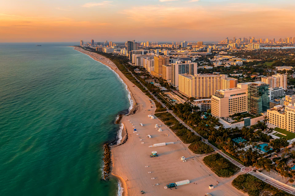Aerial view of a city coastline with tall buildings, sandy beaches, and turquoise ocean under a sunset sky.