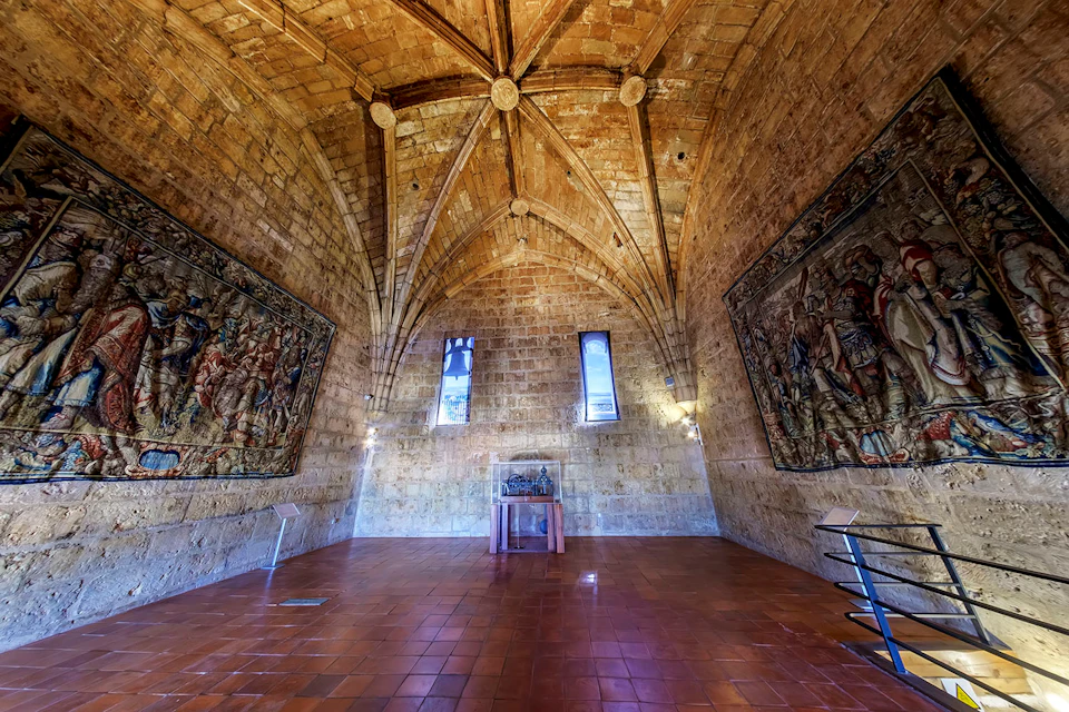 Stone-walled room with vaulted ceiling, two windows, two large tapestries, tile floor, and a small wooden table in the center.