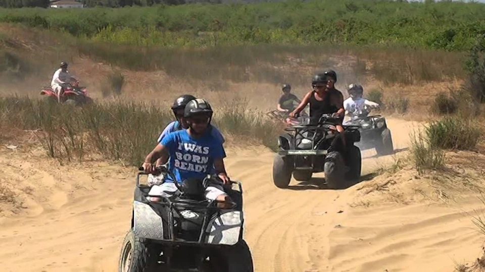 Several people ride ATVs on a dirt path, wearing helmets and casual clothes. Grass and bushes are visible in the background.