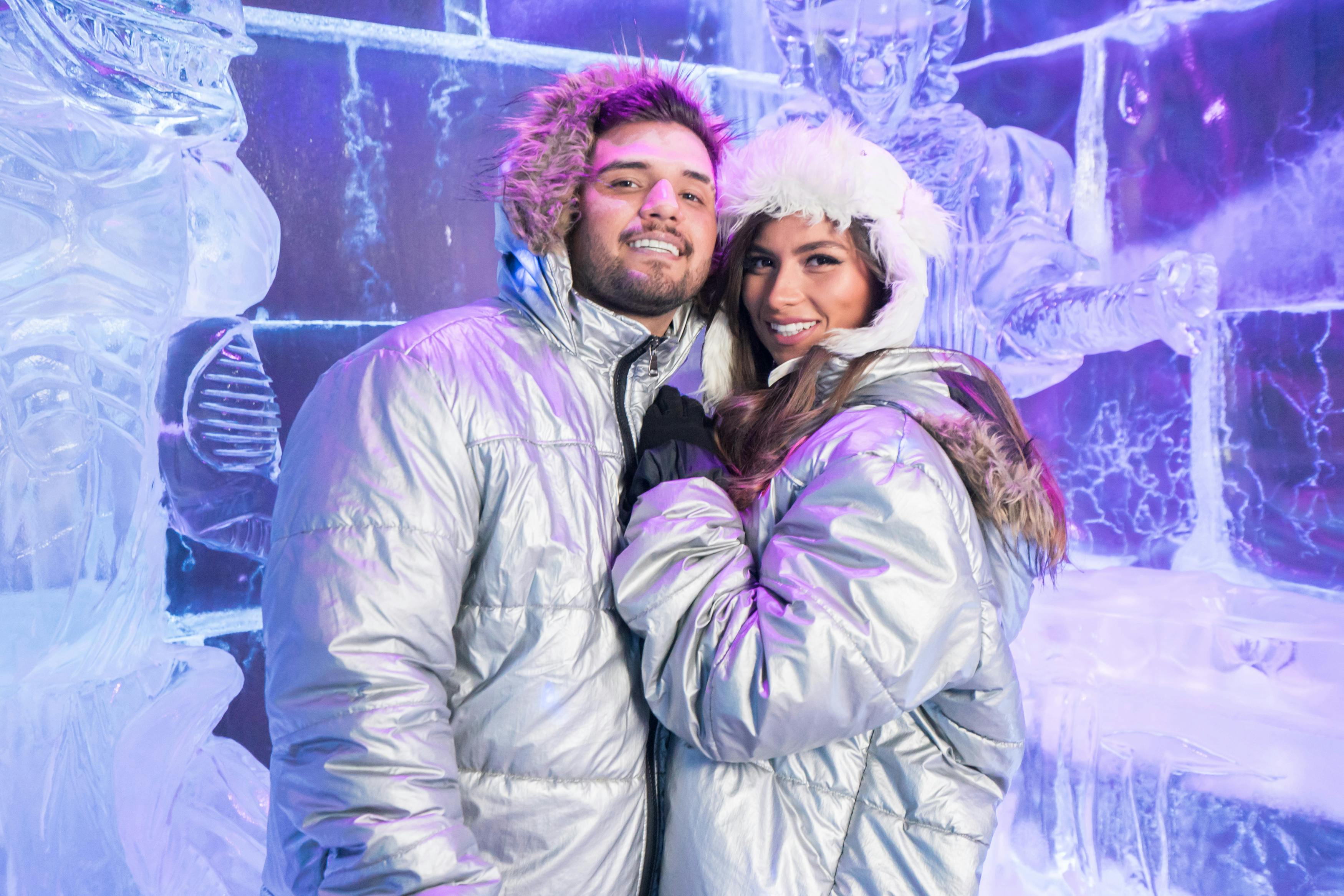 Ein Paar in silbernen Parkas steht eng beieinander, lächelnd, vor einer kunstvollen Eisskulptur mit violetter Beleuchtung.