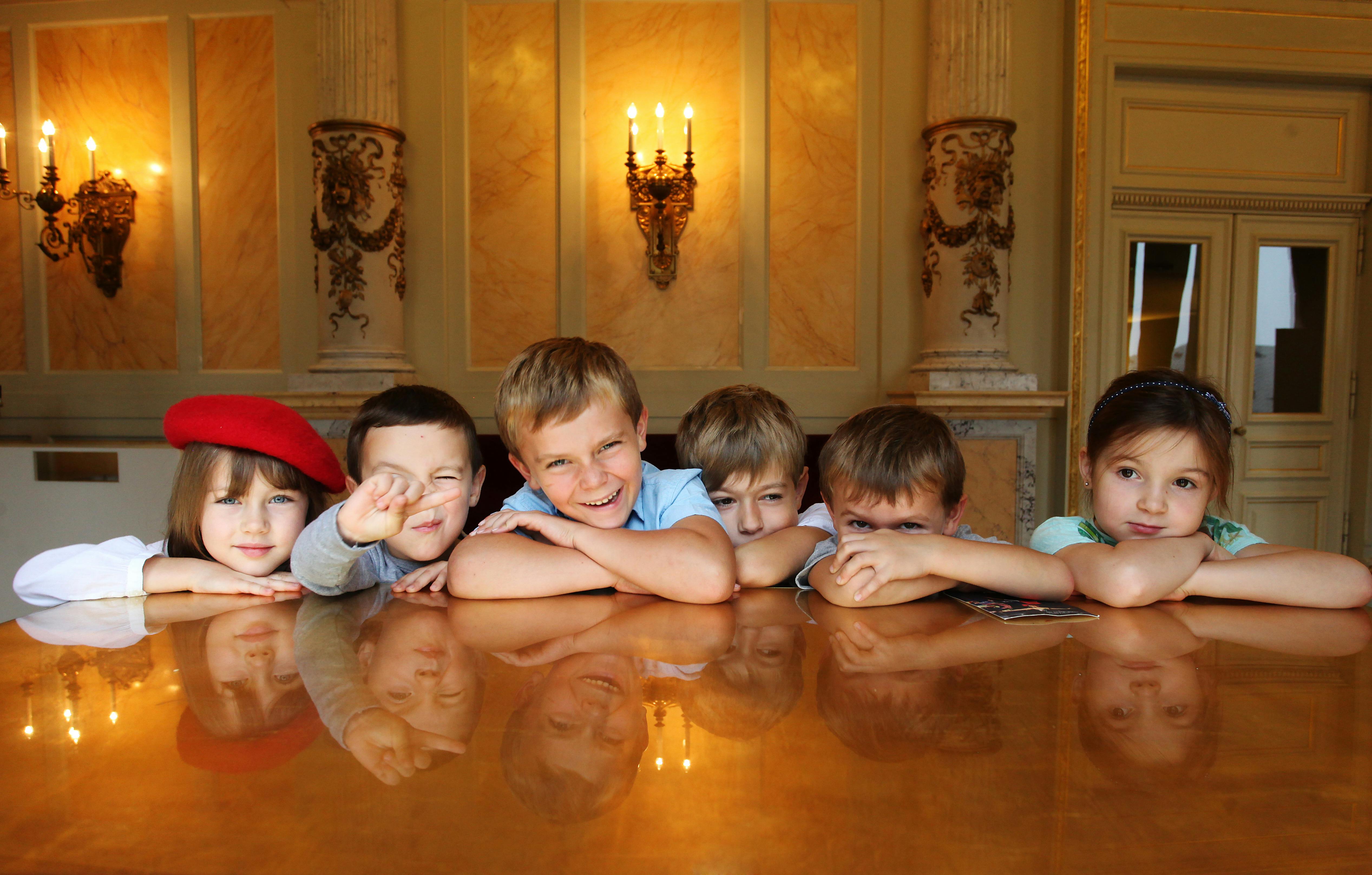 Five children lean on a polished wooden table, smiling, with ornate wall sconces and warm lighting in the background.