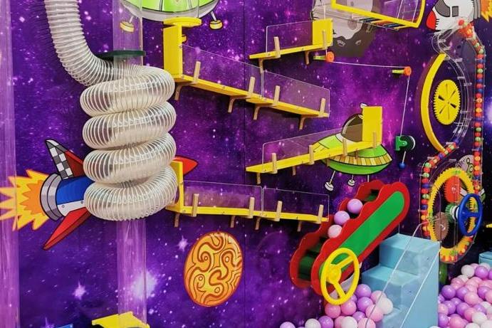 A colorful children's play area with a space theme, featuring tubes, ramps, and ball pits against a purple galaxy background.