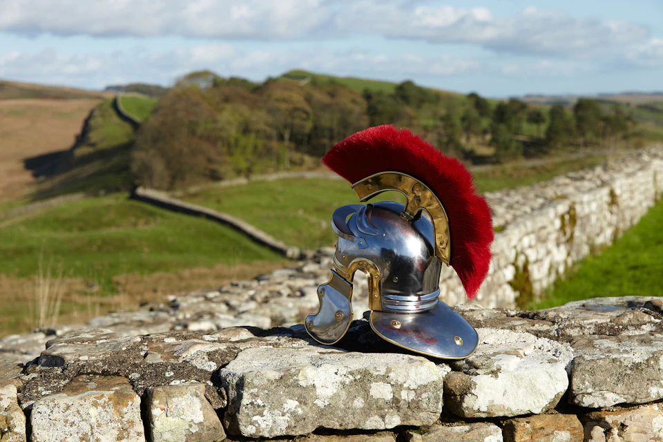 A Roman helmet with a red plume on a stone wall, with green fields and trees in the background.