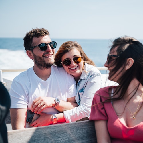 Tre persone sedute all'aperto su una barca, sorridono e indossano occhiali da sole. L'oceano e il cielo blu sono visibili sullo sfondo.