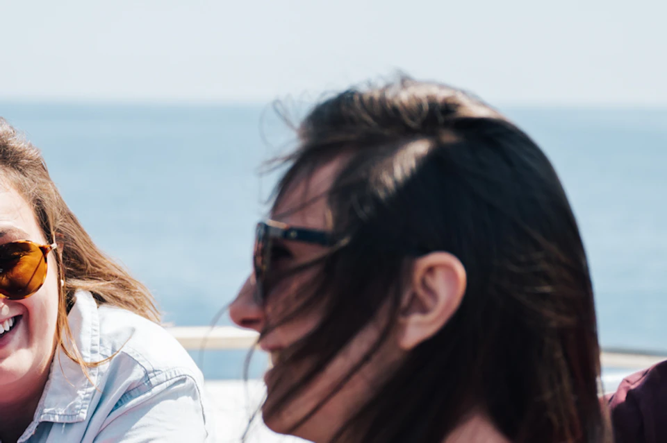 Three people sitting outside on a boat, smiling and wearing sunglasses. The ocean and blue sky are visible in the background.