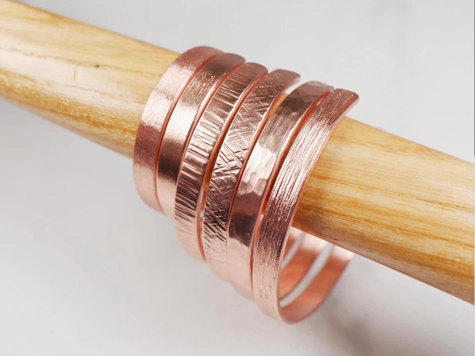 Five textured copper bangles displayed on a wooden rod.