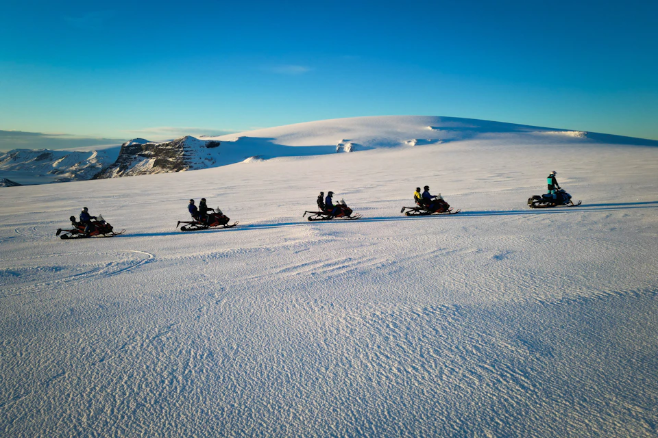 People riding snowmobiles on a snowy plain under a clear blue sky, with distant hills covered in snow in the background.