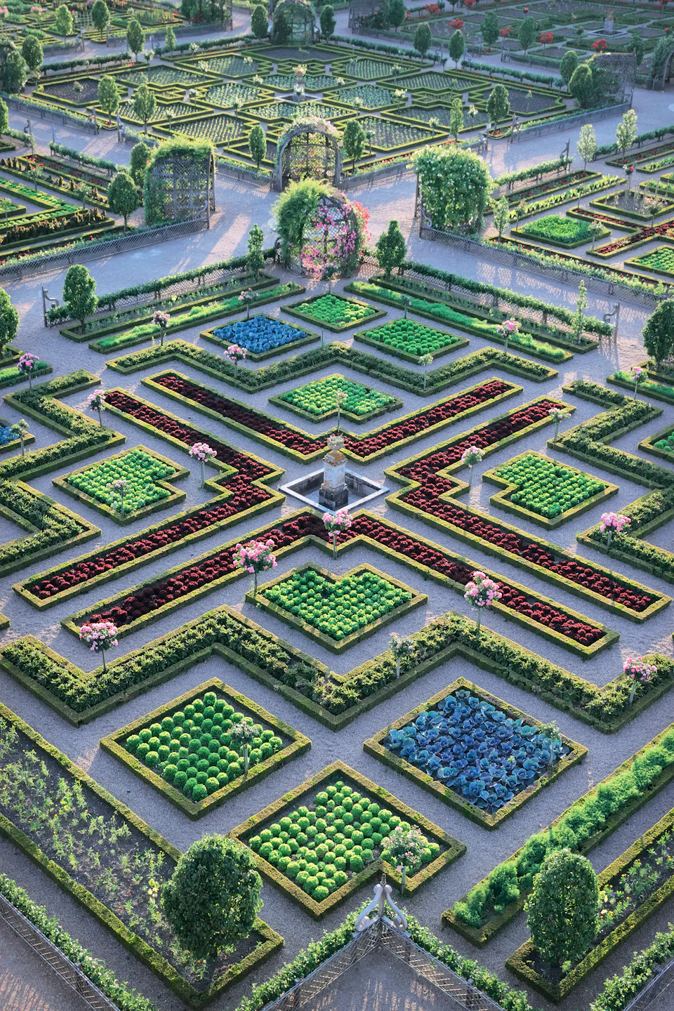 Symmetrical garden with neatly arranged flowerbeds and trimmed hedges, featuring a central fountain and pathways forming an X shape.