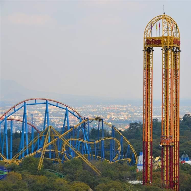 Parco divertimenti con montagne russe blu e gialle e un'alta torre di caduta rossa e gialla. Paesaggio urbano e alberi sullo sfondo.