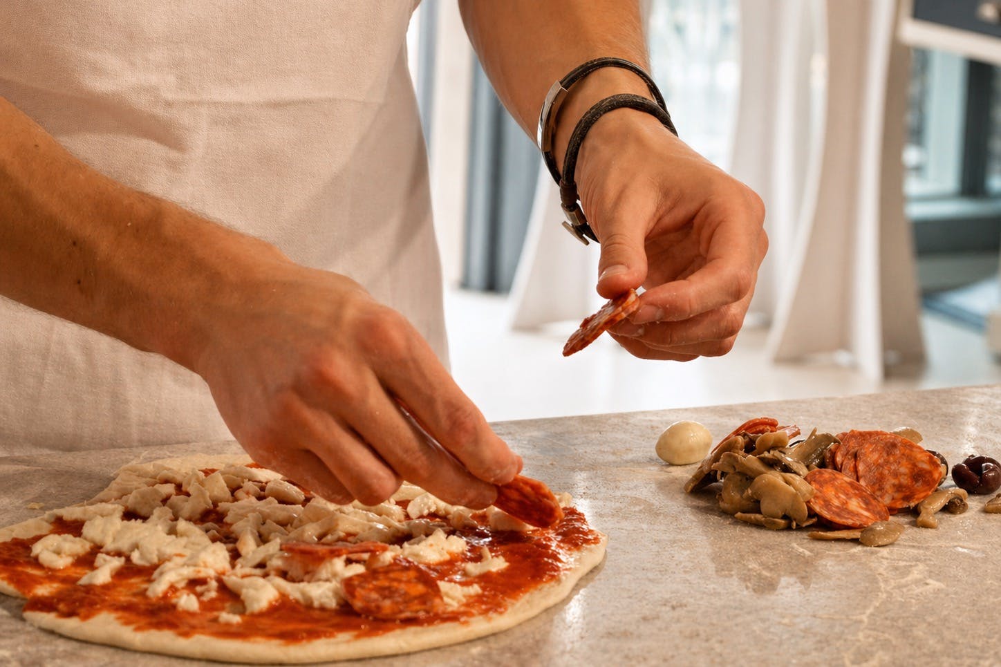 Una persona che assembla una pizza con salsa di pomodoro, formaggio e salame piccante, con i condimenti visibili su un bancone.