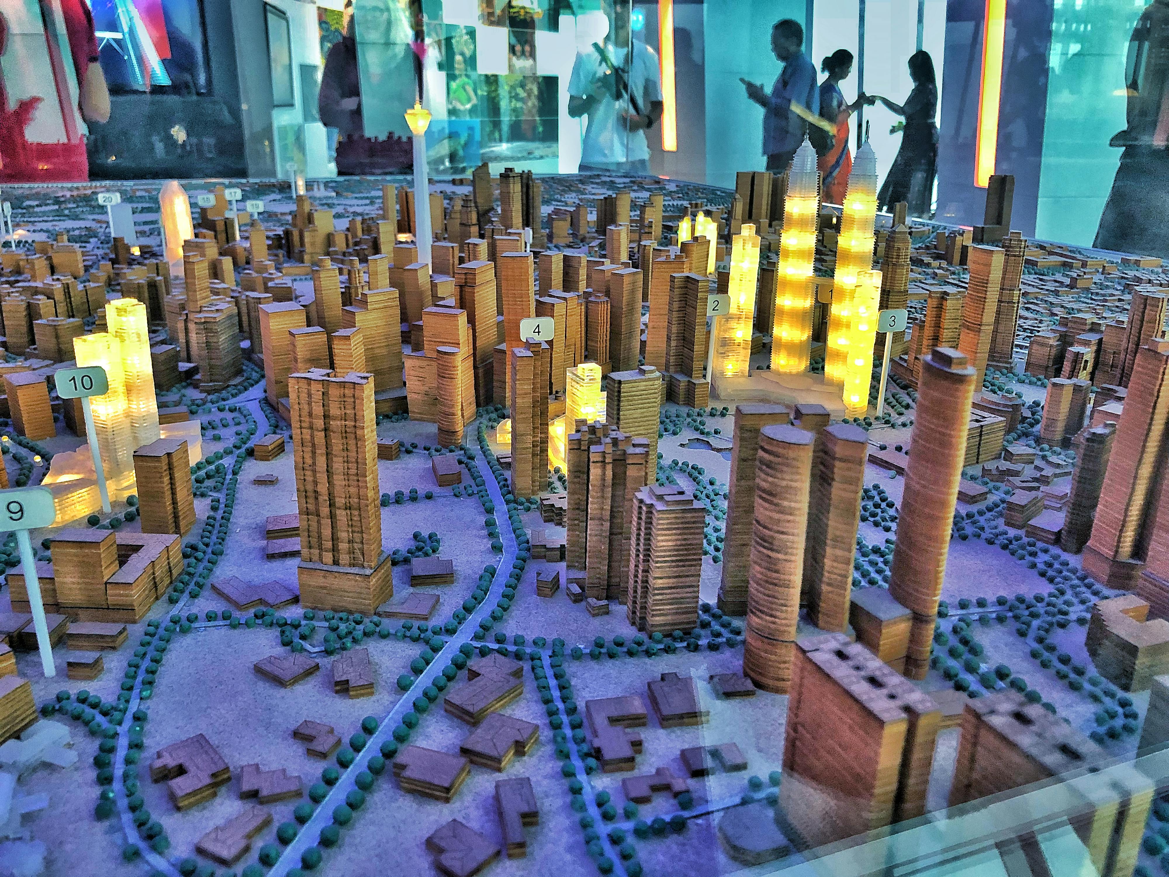 높은 건물이 있는 도시의 상세한 건축 규모 모델, 일부 조명. 배경에서 관찰하는 사람들.