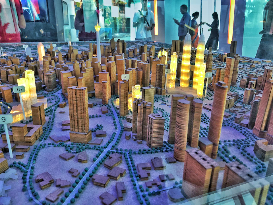 Detailed architectural scale model of a city with tall buildings, some illuminated. People observing in the background.