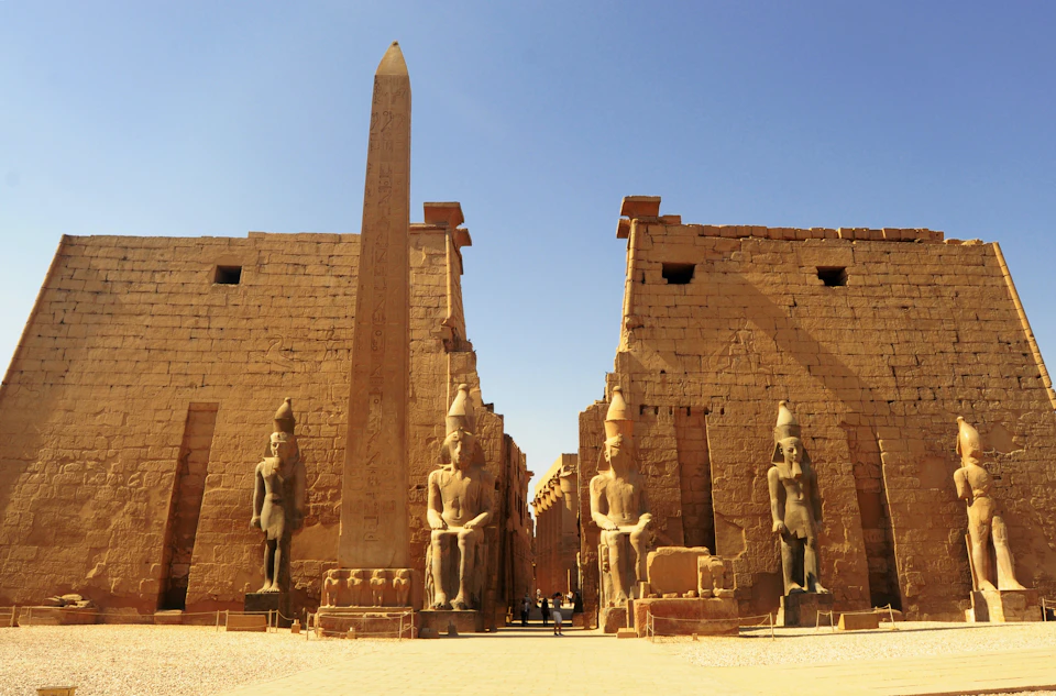 Ancient Egyptian temple entrance with large statues, an obelisk, and tourists walking under a clear blue sky.