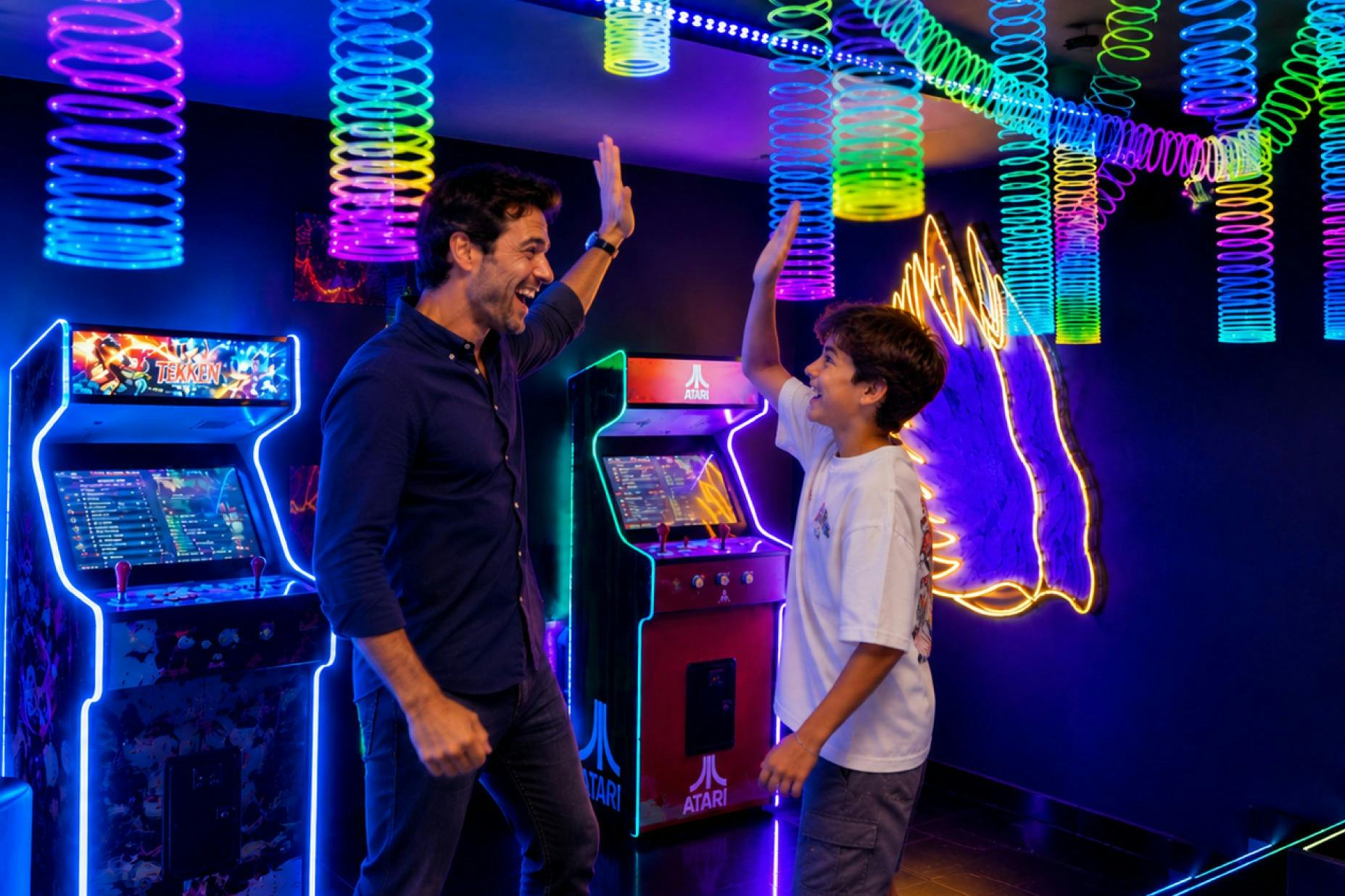 An adult and a child give a high-five in a vibrant arcade with neon lights and retro Atari game machines.