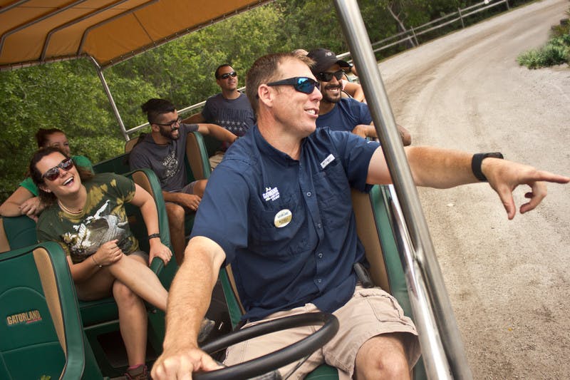 A group of people, including a smiling driver in the foreground, enjoys a ride on an open-air vehicle with "Gatorland" seats.