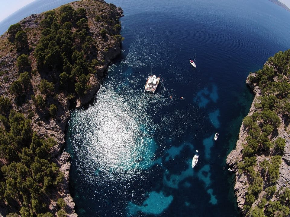 Vue aérienne de bateaux sur une mer bleue claire entre des falaises rocheuses et boisées, la lumière du soleil se reflétant sur la surface de l'eau.
