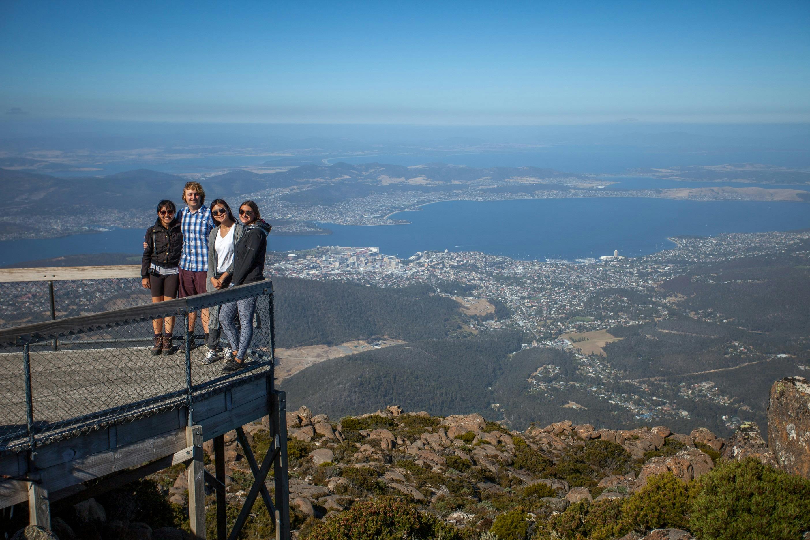 Four people standing on a wooden platform overlooking a cityscape with water and mountains in the background.