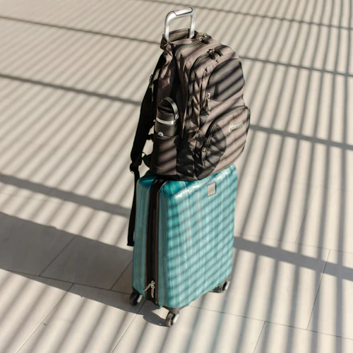 En blågrön resväska med hårt skal och en svart ryggsäck placerad på handtaget, stående på ett kaklat golv med skuggmönster.