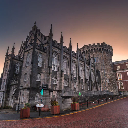 A historic stone building with pointed arches and a round tower, under a dusk sky, flanked by colorful buildings and potted plants.