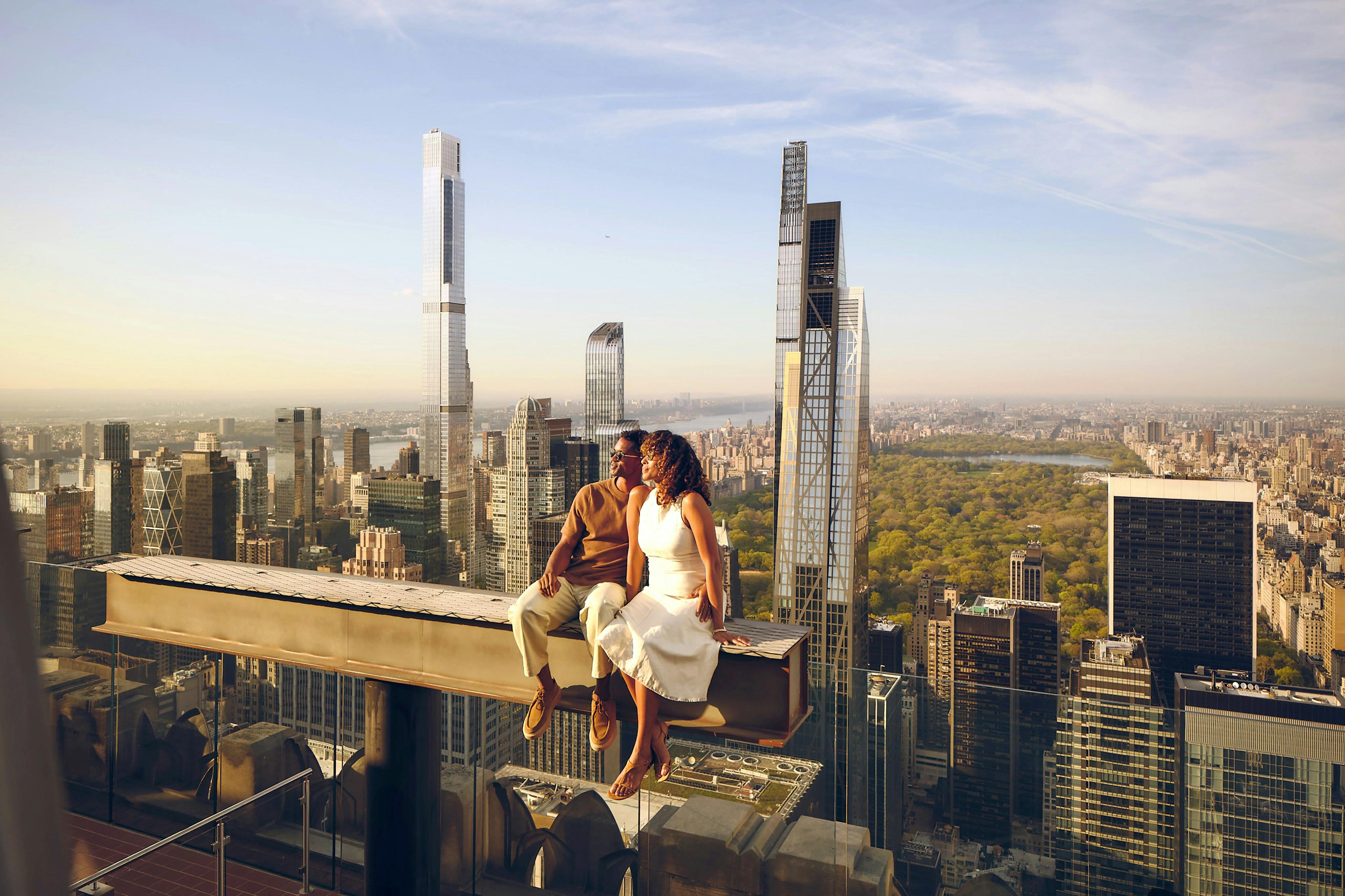 Una parella s'asseu en una biga de metall sobre l'horitzó de la ciutat amb edificis alts i un gran parc al fons.