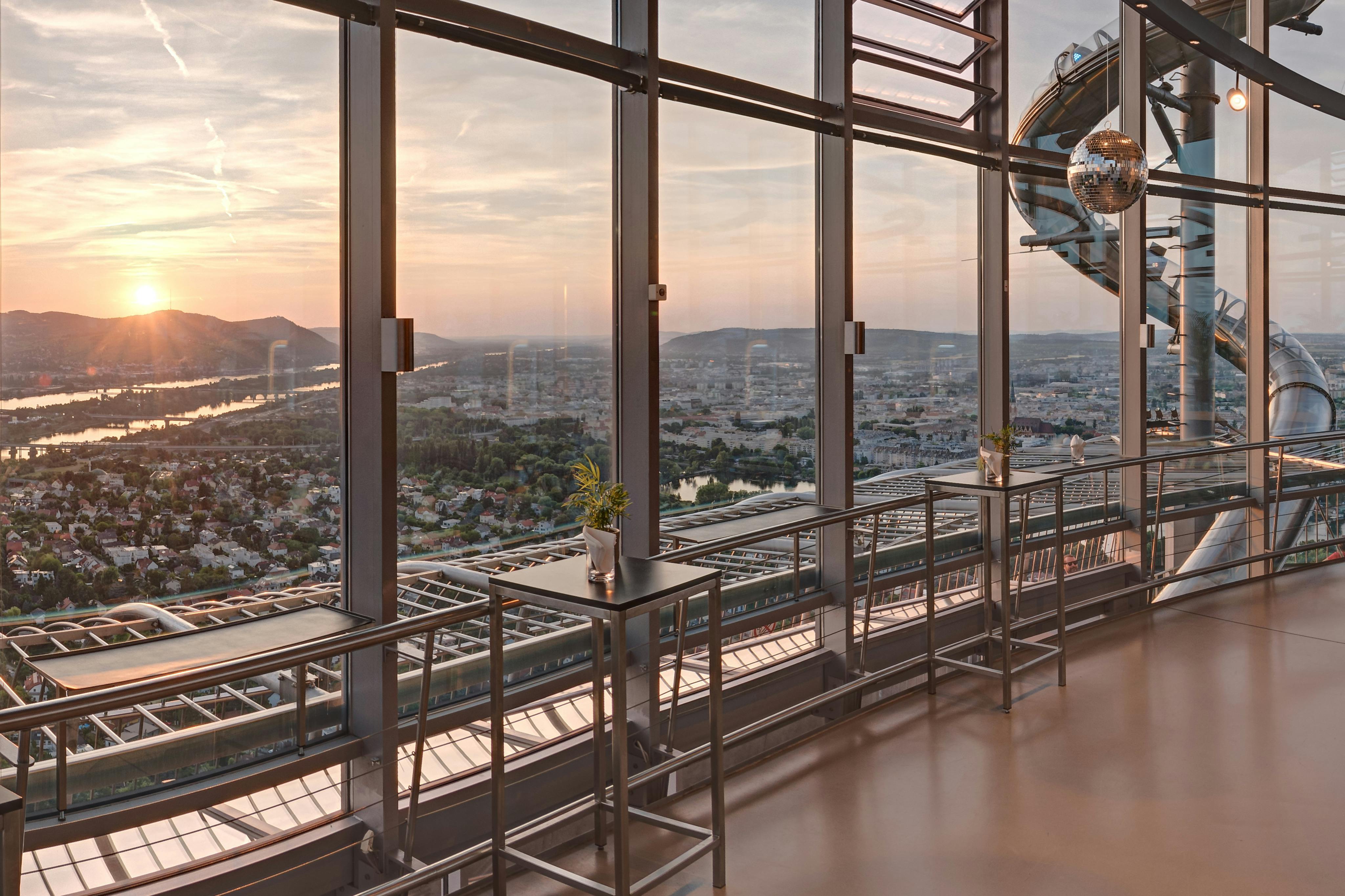 A modern interior with tall windows overlooking a cityscape at sunset, featuring high tables and stools near the windows.
