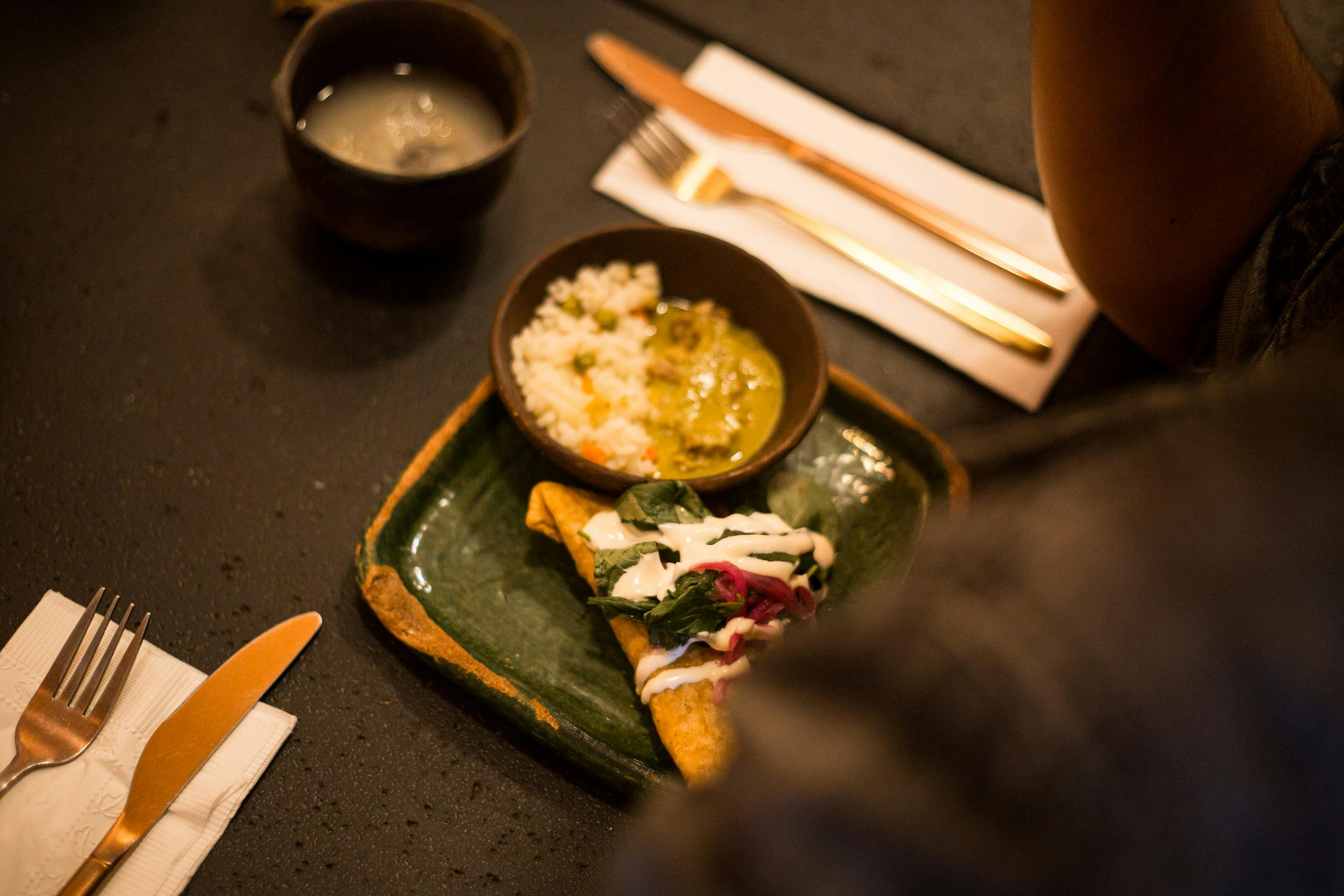 Un piatto con un taco condito con verdure e salsa, una ciotola di riso e curry e una tazza di bibita su un tavolo scuro.