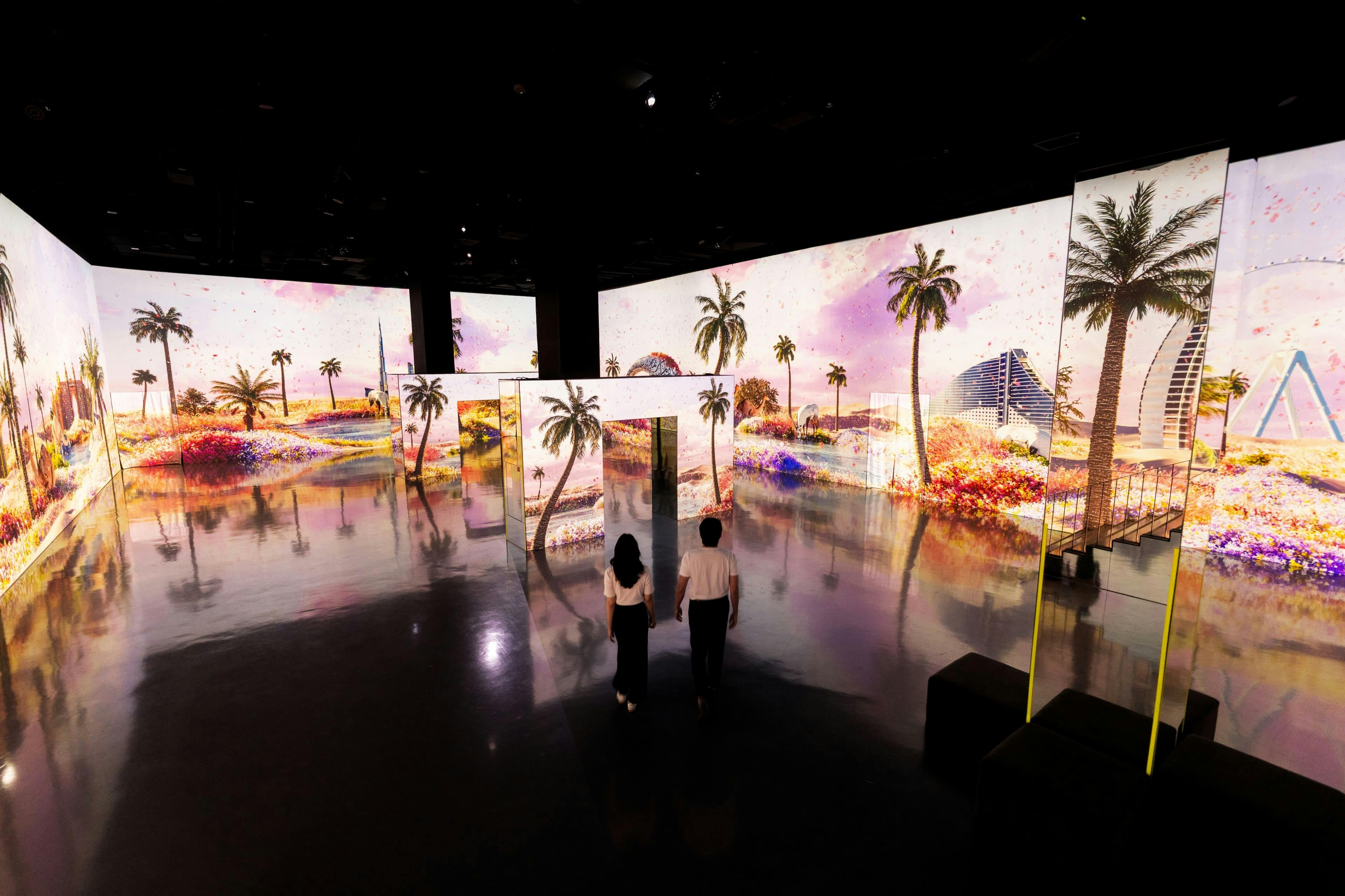 Dwie osoby przechodzą przez pokój z dużymi ekranami przedstawiającymi żywy, tropikalny krajobraz z palmami i kolorowymi kwiatami.