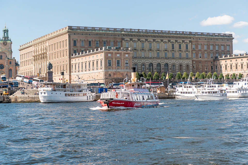 A red tour boat and several white boats docked by a large, historic building along a city waterfront on a clear day.