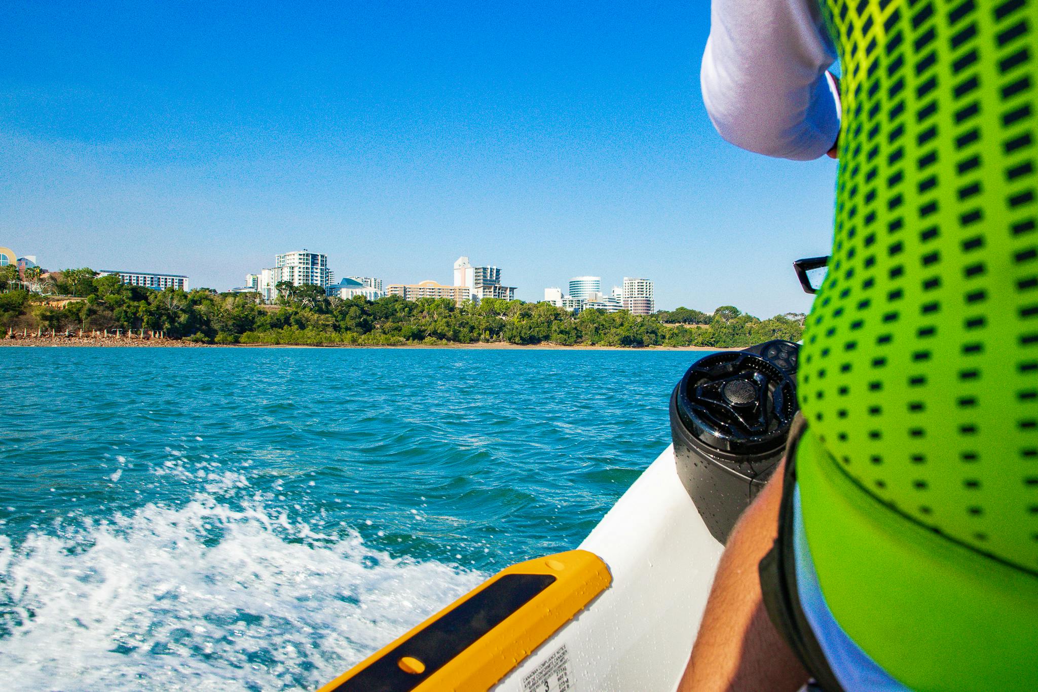 Ένα άτομο σε μια βάρκα με ένα πράσινο γιλέκο στο νερό κοντά σε ένα αστικό τοπίο κτιρίων κάτω από έναν καταγάλανο ουρανό.