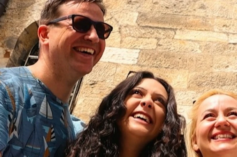 A group of five smiling people standing in front of a historic stone building with arched windows.