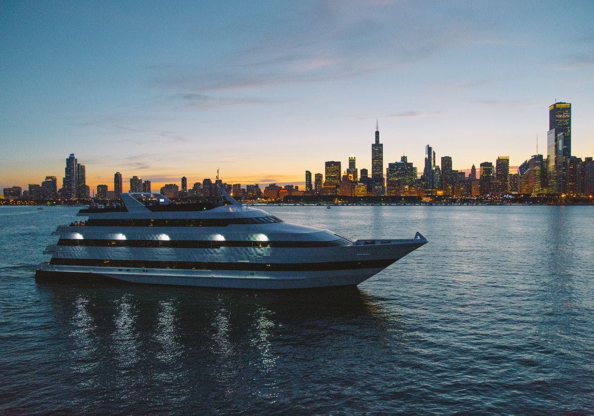 A large yacht is docked on calm water at sunset, with a city skyline featuring tall buildings in the background.